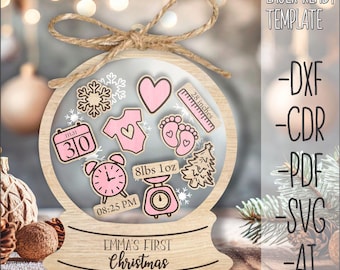 May include: A laser cut wooden ornament shaped like a snow globe with a pink and white design. The ornament includes a calendar with the date "May 30", a clock with the time "08:25 PM", a baby onesie, a baby footprint, a scale with the weight "8lbs 1oz", a ruler with the measurement "28 inches", a Christmas tree, and a heart. The ornament also includes the text "Emma's First Christmas".