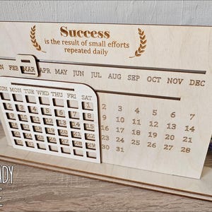 K&ouml;nnte beinhalten: Lasergeschnittenes Holz-Desktop-Kalender-Template mit einem monatlichen Kalenderraster und einem motivierenden Zitat: "Erfolg ist das Ergebnis kleiner Anstrengungen, die t&auml;glich wiederholt werden".