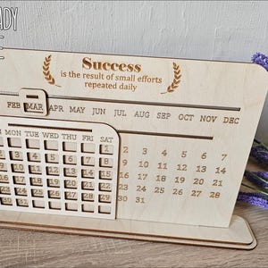 K&ouml;nnte beinhalten: Lasergeschnittene Holz-Desktop-Kalender-Vorlage mit einem monatlichen Kalenderraster und einem motivierenden Zitat: "Erfolg ist das Ergebnis kleiner Anstrengungen, die t&auml;glich wiederholt werden".