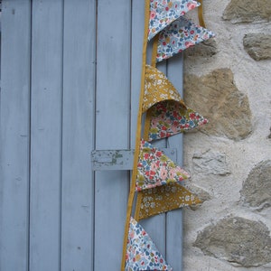 May include: A string of fabric bunting flags with a floral pattern in shades of blue, pink, and white. The flags are attached to a yellow ribbon and hang against a blue wooden door and a stone wall.