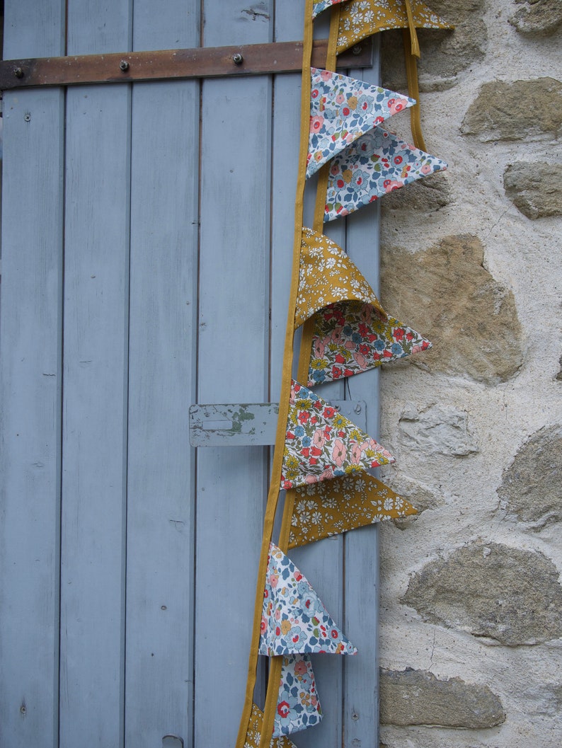 May include: A string of fabric bunting flags with a floral pattern in shades of blue, pink, and white. The flags are attached to a yellow ribbon and hang against a blue wooden door and a stone wall.