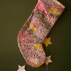 May include: A patchwork Christmas stocking constructed from various floral patterned fabric strips in shades of pink, yellow, red, and purple. The stocking features a solid cream-coloured cuff at the top and is hanging from a white wall hook.