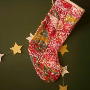 May include: A patchwork Christmas stocking constructed from various fabric scraps featuring red, pink, green, and yellow floral and geometric patterns. The stocking has a cream-coloured fabric cuff at the top and is hanging from a white wall hook.