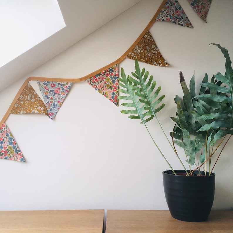 May include: A floral fabric banner with yellow, blue, and pink flowers hangs on a white wall. A potted green plant sits on a wooden table below the banner.