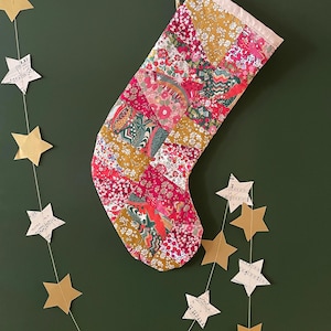 May include: A quilted Christmas stocking featuring a patchwork design of various floral and geometric fabric patterns in shades of red, pink, mustard yellow, and green, finished with a plain cream-colored fabric cuff at the top opening.