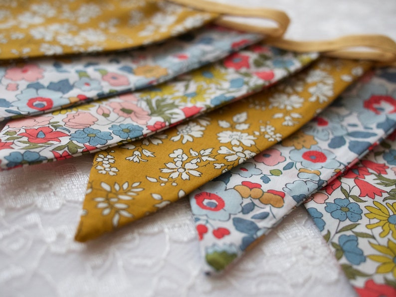 May include: A bunting banner with a yellow background and a floral pattern in shades of blue, pink, and red. The bunting is made of fabric and has a string for hanging.