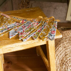 May include: A string of fabric bunting flags with a floral pattern in shades of blue, pink, and yellow. The flags are hanging on a wooden table with a woven basket in the background.
