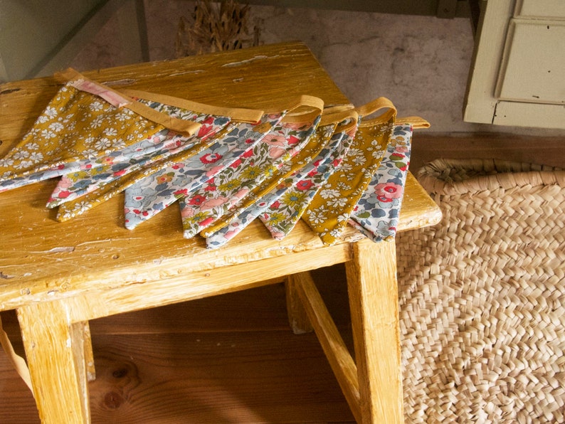 May include: A string of fabric bunting flags with a floral pattern in shades of blue, pink, and yellow. The flags are hanging on a wooden table with a woven basket in the background.