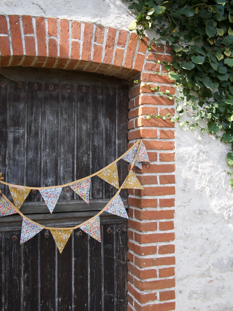 May include: A rustic wooden door with a brick archway and a floral patterned bunting hanging above it. The bunting is made of fabric triangles with a yellow trim. The door is weathered and has a dark brown finish.