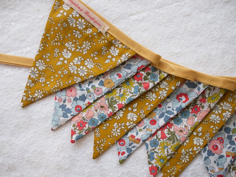 May include: A bunting banner with a yellow background and a floral pattern. The banner is made of fabric and has a white trim. The banner says " Bunting Boutique" on one of the flags.