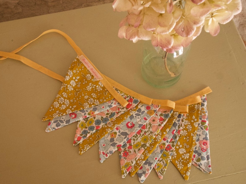 May include: A yellow and white floral fabric bunting with a yellow ribbon. The bunting is made of triangular flags with a variety of floral patterns.