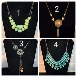 May include: Four different necklaces, each with a unique design. The first necklace is a light green statement necklace with large, faceted stones. The second necklace is a gold-toned necklace with a large, round pendant and a long, pointed crystal. The third necklace is a silver-toned necklace with a large, black flower pendant and a long, beaded tassel. The fourth necklace is a silver-toned necklace with a large, multi-layered pendant made of turquoise and green beads.