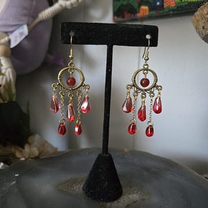 Peut inclure: Une paire de boucles d'oreilles chandelier avec des cadres circulaires en métal doré vieilli et des détails en filigrane. Chaque boucle est ornée d'une perle rouge centrale et de plusieurs breloques en verre rouge translucide.