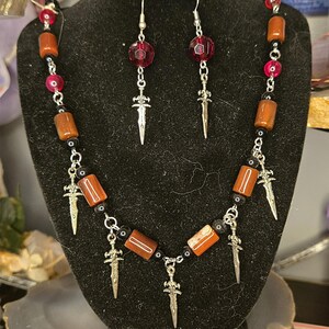 May include: A jewelry set consisting of a necklace and matching earrings. The necklace features cylindrical amber-colored beads, small black beads, and silver-toned sword charms. The earrings include a faceted dark red bead and a silver sword charm.