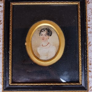 Op de afbeelding: Een ingelijst portret van een vrouw met donker haar en een witte jurk. Het portret is geplaatst in een gouden ovale lijst en omgeven door een zwarte lijst met gouden accenten.