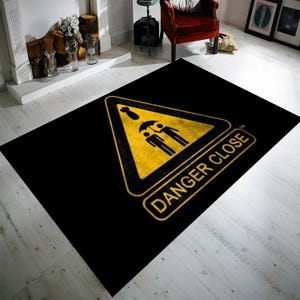 May include: Black and yellow rug with a warning sign that says "Danger Close" and a graphic of three people under an umbrella.