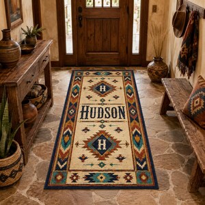 May include: A rectangular runner rug featuring a cream-colored center with the text HUDSON in dark blue letters, flanked by two diamond-shaped geometric patterns containing the letter H, all bordered by a multi-colored southwestern-style motif.