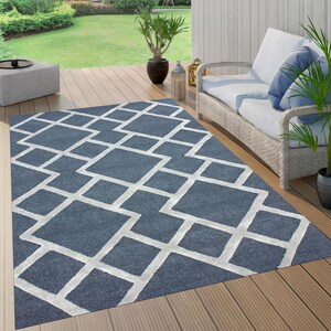 May include: A blue and white geometric outdoor rug with a modern design. The rug is laid out on a wooden deck with a white wicker sofa and a potted plant in the background.