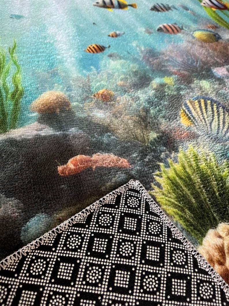 May include: A rectangular floor mat featuring a vibrant underwater coral reef scene with various colorful tropical fish, green seaweed, and textured rocks. The underside of the mat is black with a white geometric grid pattern and stitched edges.