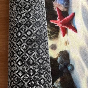 May include: A rectangular mat featuring a beach scene with a bright red starfish, blue coral, and dark rocks on white sand. The underside of the mat is black with a repeating white geometric pattern of small dots forming squares and diamonds.