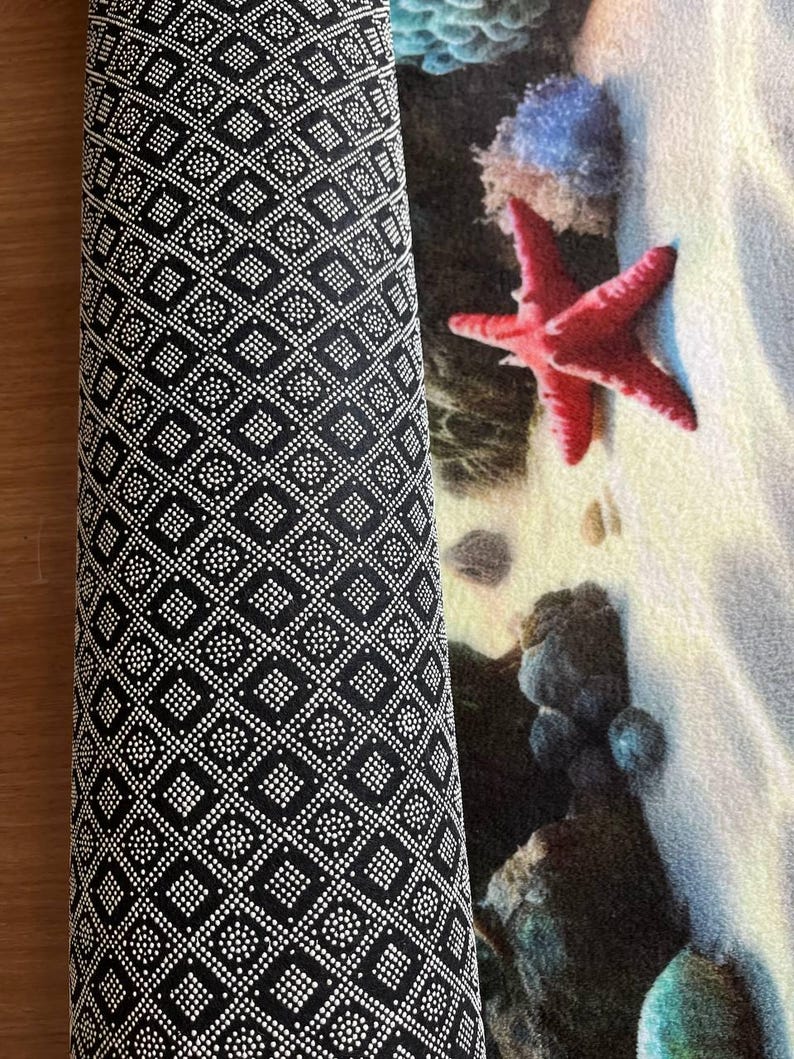 May include: A rectangular mat featuring a beach scene with a bright red starfish, blue coral, and dark rocks on white sand. The underside of the mat is black with a repeating white geometric pattern of small dots forming squares and diamonds.