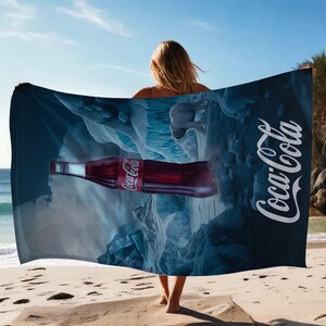 May include: A blue beach towel featuring a Coca-Cola logo and a polar bear walking on a snowy mountain scene. A bottle of Coca-Cola is lying on the snow.
