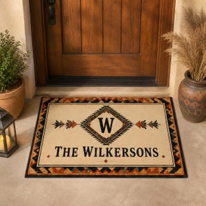 May include: A rectangular doormat with a cream-colored center featuring a central diamond containing the letter W, flanked by arrow motifs. The text THE WILKERSONS is printed below. The border has a geometric pattern in black, orange, and blue.