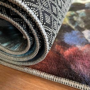 May include: A rolled area rug featuring a black underside with a white geometric diamond pattern and non-slip rubberized dots. The top surface displays a blurred, multi-colored abstract design in shades of deep red, blue, and dark tones.