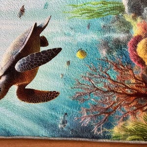 May include: A rectangular rug featuring a detailed underwater scene with a brown sea turtle swimming toward a vibrant coral reef. The reef displays shades of red, yellow, and green, set against a textured blue ocean background with small fish.