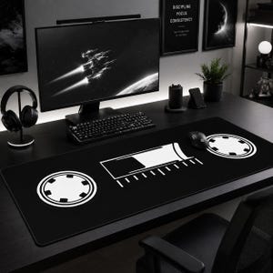 Può includere: Tappetino da scrivania rettangolare nero con un design grafico bianco. Il design include una forma rettangolare centrale con linee parallele e due forme circolari con un motivo a stella. Un mouse per computer è sul tappetino.