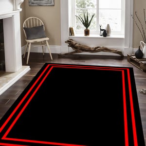 May include: Black area rug with a double red border. The rug is in a living room setting with a fireplace and a window.