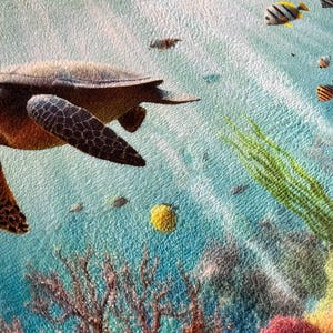 May include: A textured fabric print featuring an underwater scene with a brown sea turtle swimming through light blue water. The scene includes small colorful tropical fish, green seaweed, and brown coral formations at the bottom of the frame.