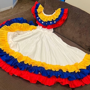 May include: A white dress with layers of red, yellow, and blue frills. The top has a frilled collar with yellow, blue, and red accents and white stars. The dress is likely a traditional costume.