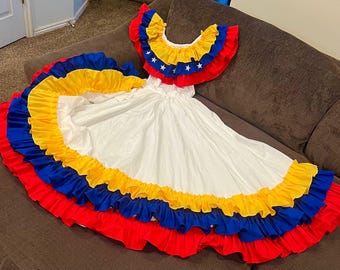 May include: A white dress with layers of red, yellow, and blue ruffles. The top has a ruffled collar with yellow, blue, and red accents and white stars. The dress is likely a traditional costume.