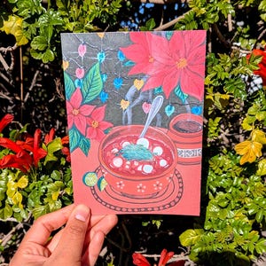 May include: A red greeting card featuring a vibrant illustration of a bowl of pozole, a traditional Mexican soup, with red poinsettias and decorative lights in the background. A small cup of coffee is also depicted. The card is held against a backdrop of green foliage and red flowers.