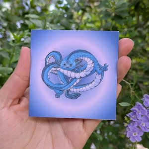 May include: Square art print with a blue dragon illustration. The dragon is in a stylized, intertwined pose against a light blue and purple gradient. The artwork is held in a hand, with a blurred green and purple floral background.