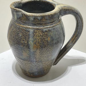 Könnte beinhalten: Eine handgefertigte Keramikkrug mit einem großen Griff. Der Krug hat eine dunkelblaue und braune, gesprenkelte Glasur mit einer strukturierten Oberfläche. Der Rand und der Griff haben eine hellere, beige Farbe.