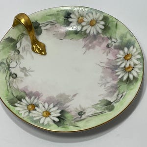 Puede incluir: Un plato redondo de porcelana pintado a mano con un asa dorada. El plato presenta un interior blanco con un diseño floral de margaritas y detalles en verde y rosa. El borde está adornado con oro.