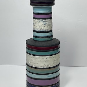 May include: A decorative vase with a striking striped pattern. The vase is predominantly cylindrical, with bands of blue, purple, white, teal, and black. The top and base are dark, and the textured white band adds visual interest.