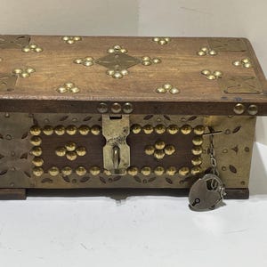 May include: A wooden chest with brass accents and a lock. The chest has a rectangular shape with a wooden top and sides adorned with brass studs and decorative metal plates. The lock is attached with a chain.