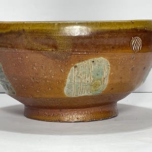 May include: A handcrafted ceramic bowl with a wide, rounded shape. The bowl features a warm color palette of brown, tan, and green, with a textured surface and decorative accents. The bowl is approximately 6 inches in diameter and 4 inches tall.