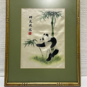 May include: Framed artwork featuring a panda bear in black and white, reaching for bamboo. The artwork is set against a green background and framed in a gold-colored bamboo-style frame. The artwork includes Chinese characters.
