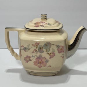 May include: Cream-colored vintage teapot with a square body and a matching lid. The teapot features a floral pattern in pink and blue hues. The spout and handle are silver-colored, adding a touch of elegance to this classic tea accessory.