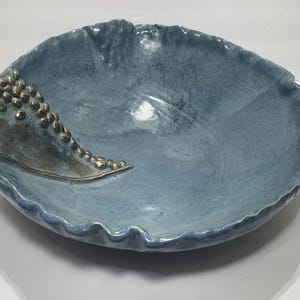 Pode incluir: Uma grande tigela de cerâmica feita à mão em um tom azul suave. A tigela apresenta uma borda texturizada e ondulada e um detalhe decorativo de contas prateadas em relevo e uma linha curva. O interior da tigela tem um acabamento brilhante.
