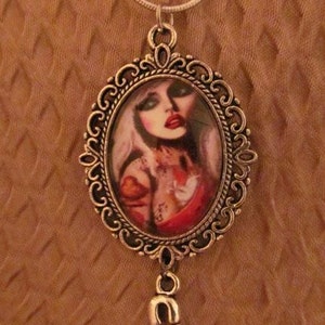 Op de afbeelding: Zilverkleurige hangerketting met een ovale cameo met een vrouwenportret, rode lippen en tatoeages. De hanger is omlijst door een sierlijke, filigraan-achtige rand en bevat een veiligheidsspeld-bedel. Zilveren ketting.
