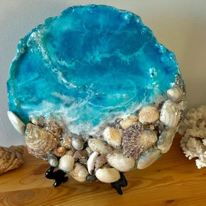 May include: A round resin art piece depicting a blue ocean scene with white foamy waves and a beach shore covered in seashells. The piece is mounted on a black stand.