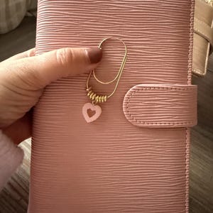 Op de afbeelding: Een roze, getextureerde leren planner met een goudkleurige paperclip en een roze hartbedel. De planner heeft een klepsluiting en wordt door een hand vastgehouden. Het algemene thema is briefpapier en organisatie.