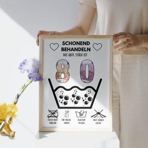 Op de afbeelding: Een witte geframed poster met zwarte tekst die "Schonend Behandeln Das Gute St&uuml;ck Ist 80" luidt. De poster heeft een zwart-witte illustratie van een wasbak met bubbels en symbolen voor wasinstructies.