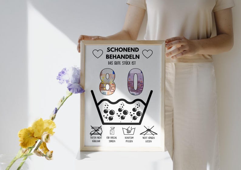 Op de afbeelding: Een witte geframed poster met zwarte tekst die "Schonend Behandeln Das Gute St&uuml;ck Ist 80" luidt. De poster heeft een zwart-witte illustratie van een wasbak met bubbels en symbolen voor wasinstructies.