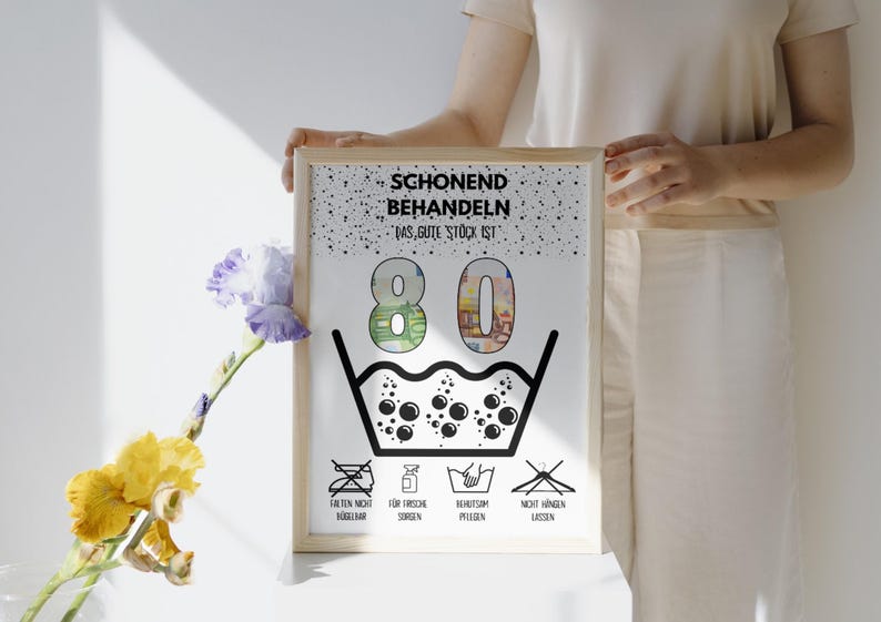 Op de afbeelding: Een witte kaart met zwarte tekst die "Schonend Behandeln Das gute Stock ist 80" zegt met een zwart-witte illustratie van een wasbak met bubbels. De kaart heeft vier zwart-witte pictogrammen onder de bak die "Talten Nicht Heiss", "For Frische Sorgen", "Behutsam Pflegen" en "Nicht Hangen Lassen" zeggen.